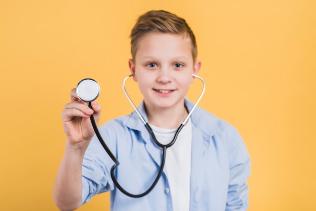 Portrait of a smiling boy holding stethoscope towards camera standing against yellow background