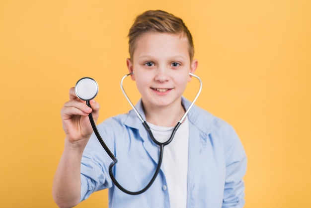 Портрет улыбающегося мальчика, удерживая стетоскоп на камеру, стоя на желтом фоне