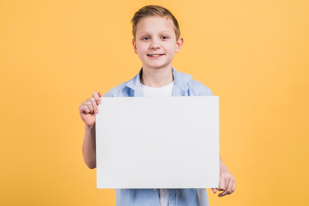 Портрет улыбающегося мальчика, глядя в камеру, показывая белый пустой плакат на желтом фоне