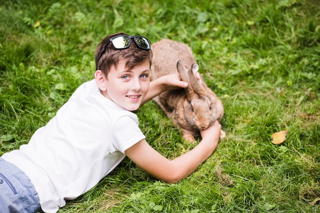 Портрет улыбающегося мальчика, лежащего на зеленой траве, заботясь о своем кролике
