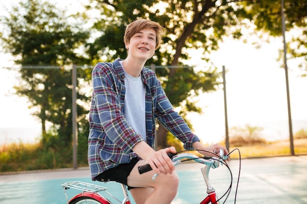 ショートパンツとカジュアルなシャツを着た笑顔の少年の肖像画公園のバスケットボールコートで自転車に乗って赤い自転車で立っている間夢のように脇を見て少年