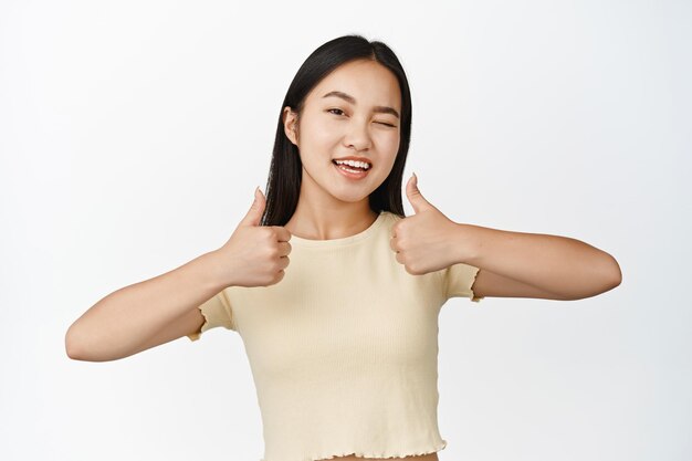 Portrait of smiling brunette asian girl showing thumbs up winking and looking at camera happy encourage you standing over white background