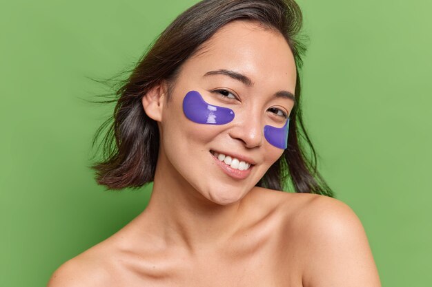 Portrait of smiling brunette Asian woman with natural beauty applies blue hydrogel patches under eyes