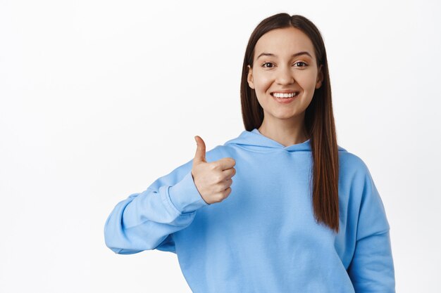 Portrait of smiling brunette female, showing thumb up, looking upbeat and satisfied, recommend smth good, praise excellent company, perfect quality, wearing hoodie.