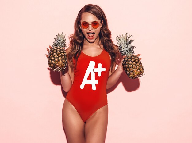 夏の赤い水着服と丸いサングラスで笑顔ブルネットの少女の肖像画。新鮮なパイナップルとセクシーな女性。ポジティブモデルのポーズ