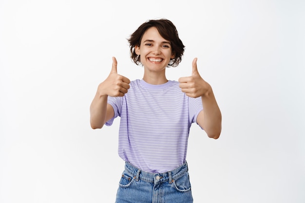 Free photo portrait of smiling brunette woman showing thumbs up, like and approve, positive feedback, praise and compliment smth good, well done gesture, standing on white.
