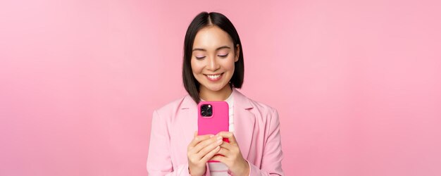 Portrait of smiling business woman asian corporate person using smartphone mobile phone application standing over pink background