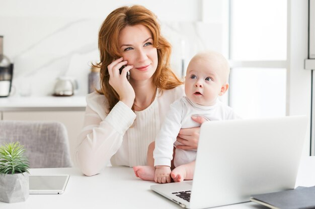 노트북을 들고 테이블에 앉아 귀여운 아기를 손에 들고 휴대폰으로 통화하는 웃고 있는 비즈니스 여성의 초상화