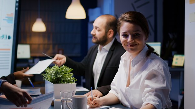 Portrait of smiling businesswoman looking into camera working overtime in business company meeting office room late at night. Workaholics diverse multi-ethnic teamwork discussing investment strategy