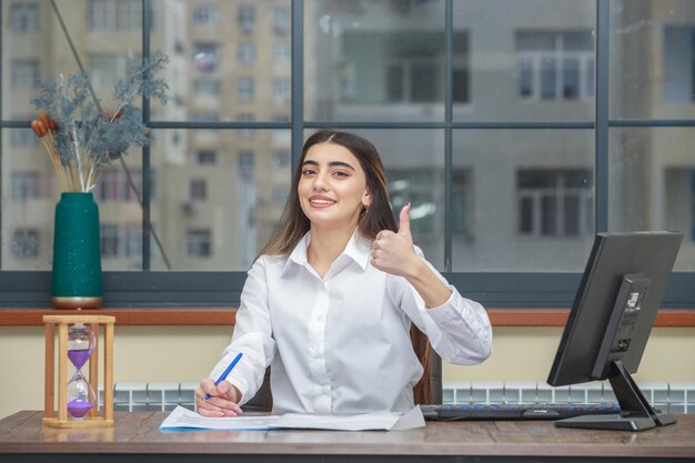 Портрет улыбающейся деловой женщины, сидящей за столом и поднимающей большой палец вверх