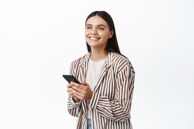 Portrait of smiling caucasian woman in trendy outfit, chatting or using app, looking happy aside, standing against white wall