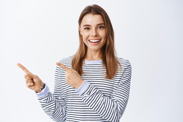 Portrait of smiling caucasian woman with blond hair, showing good deal offer, pointing fingers left at logo banner and looking at front, show direction or way, white wall