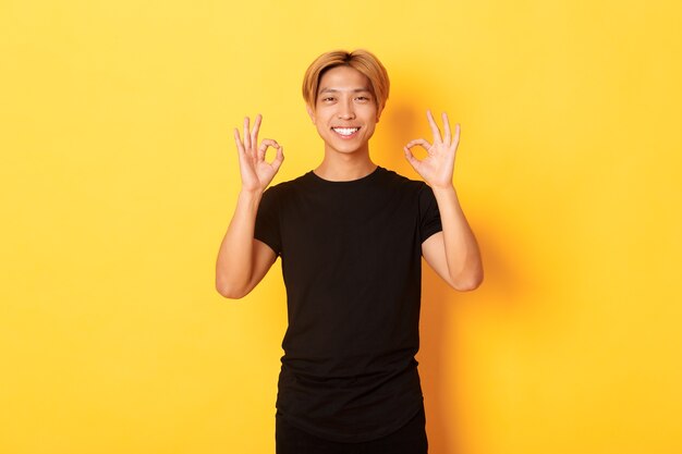 미소 자신감 아시아 남자의 초상화, 기쁘게 찾고 괜찮 제스처, 노란색 벽을 보여주는