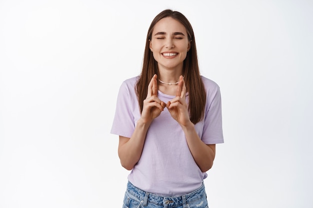 Portrait of smiling cute girl holds fingers crossed with eyes closed anticipate exam results, waiting for dream come true on white.