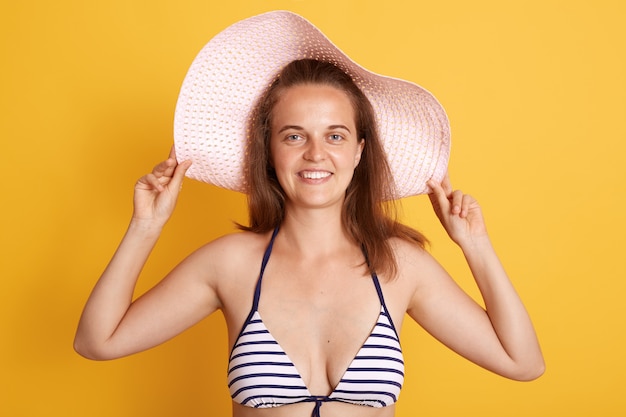Portrait of smiling dark haired woman woman wearing straw hat and striped white and black swimming suit with charming smile isolated over yellow wall.