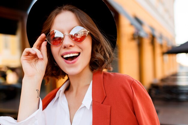 Портрет улыбающегося европейской девушки в милые оранжевые очки, куртка и черная шляпа. Осенняя мода. Уличное кафе.