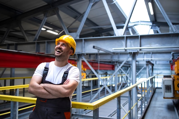Портрет улыбающегося фабричного рабочего со скрещенными руками, стоящего в производственном цехе