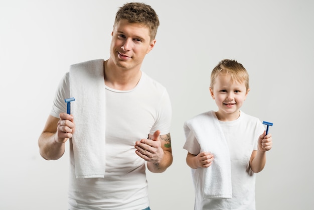 Portrait of a smiling father and son holding razor in hand looking to camera against white backdrop