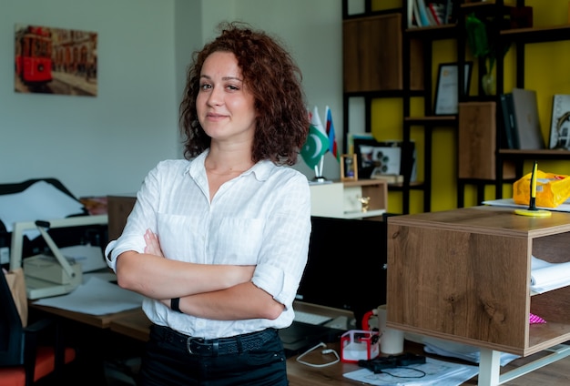 Portrait of smiling female employee standing with crossed arms looking confident wearing white shirt standing in office
