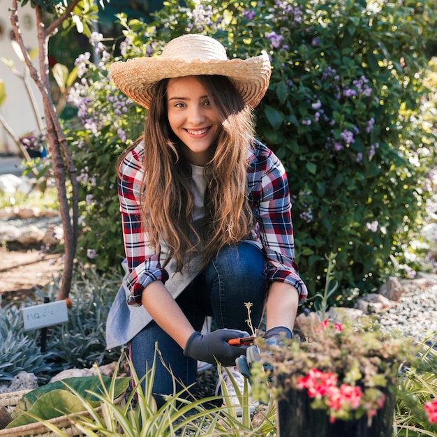 Portrait of a smiling female gardener wearing hat cutting the plants in the garden