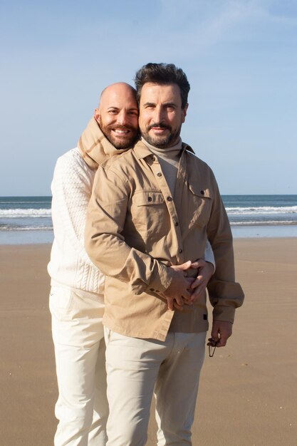 Portrait of smiling gay couple hugging on beach. Cheerful bald man keeping arms around waist of boyfriend. Gays concept