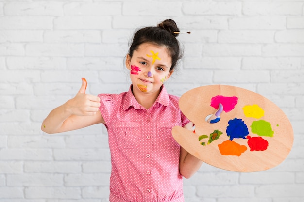 Portrait of a smiling girl holding multi colored palette showing thumb up sign standing against white brick wall