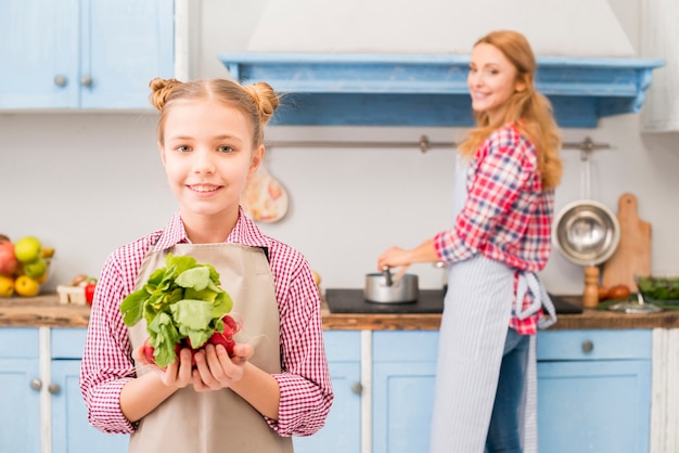 Portrait of smiling girl holding radish and spinach in hand with her mother at background