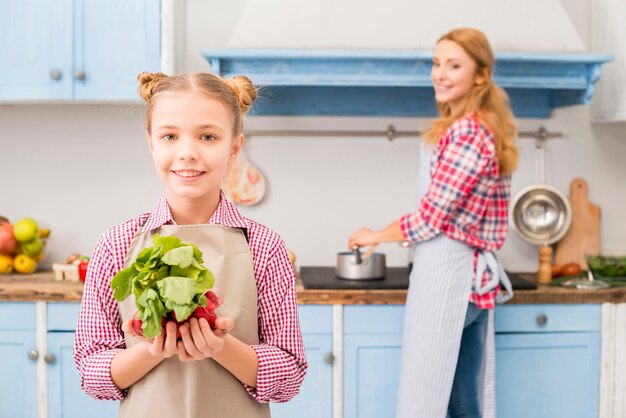 Portrait of smiling girl holding radish and spinach in hand with her mother at background