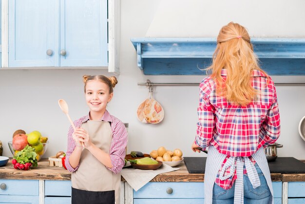 Portrait of a smiling girl holding spoon in hand and her mother cooking food in the kitchen