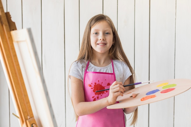 Portrait of a smiling girl holding wooden palette and paintbrush standing against white wooden wall