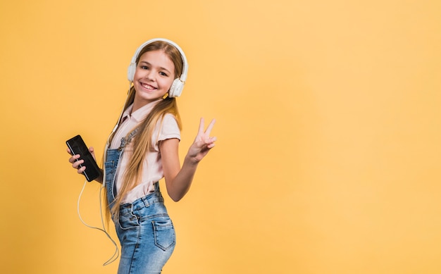 Portrait of a smiling girl listening music on white headphone gesturing against yellow backdrop