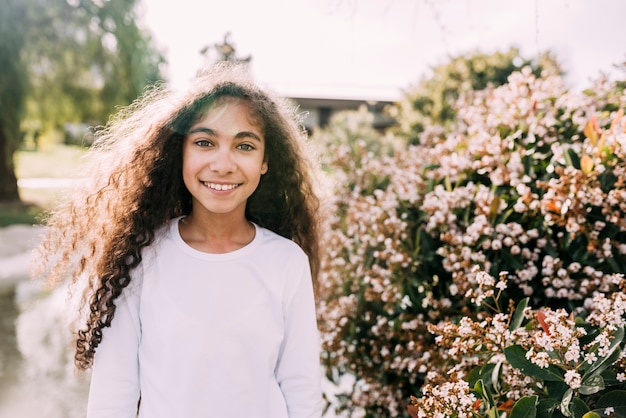 Free photo portrait of a smiling girl looking at camera standing in front of flowers plant