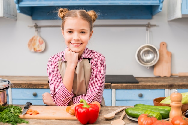 Free photo portrait of a smiling girl looking at camera standing behind the table with vegetables