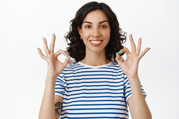 Portrait of smiling girl shows okay OK signs look satisfied recommend good company perfect quality praise good job well done standing pleased against white background