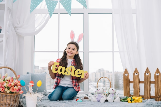 Free photo portrait of a smiling girl sitting in front of window showing yellow easter word