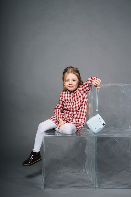 Portrait of a smiling girl sitting on transparent blocks holding retro instant camera in hand