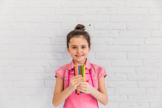 Portrait of a smiling girl standing against white brick wall holding colored pencils in hand