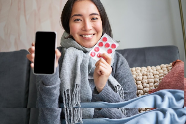 Портрет улыбающейся девушки, которая болеет дома и показывает экран мобильного телефона, рекомендуя онлайн-медицину