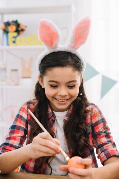 Portrait of a smiling girl wearing bunny ears painting easter egg with brush