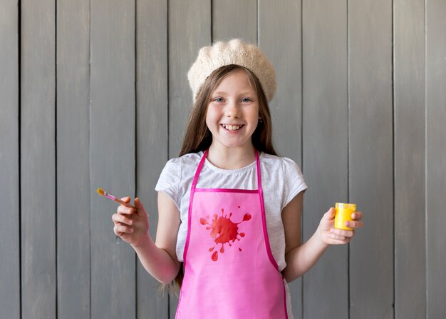 Portrait of a smiling girl wearing knit cap holding paintbrush and yellow paint bottle in hands