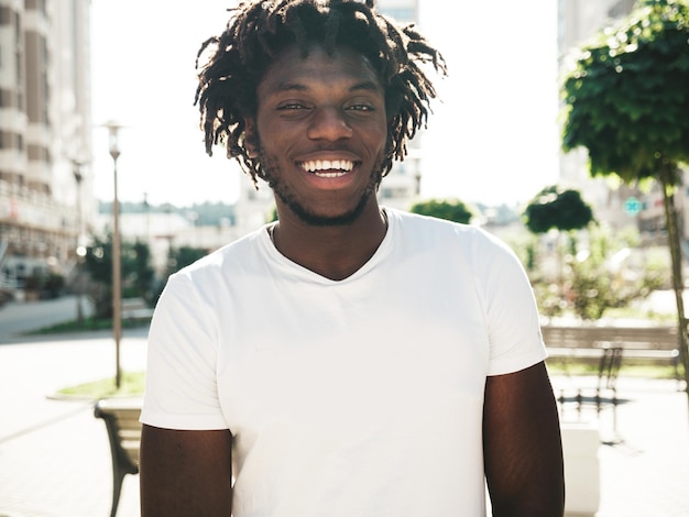 Portrait of smiling handsome hipster modelunshaven african man dressed in white summer tshirt and jeans fashion male with dreadlocks hairstyle posing on the street background