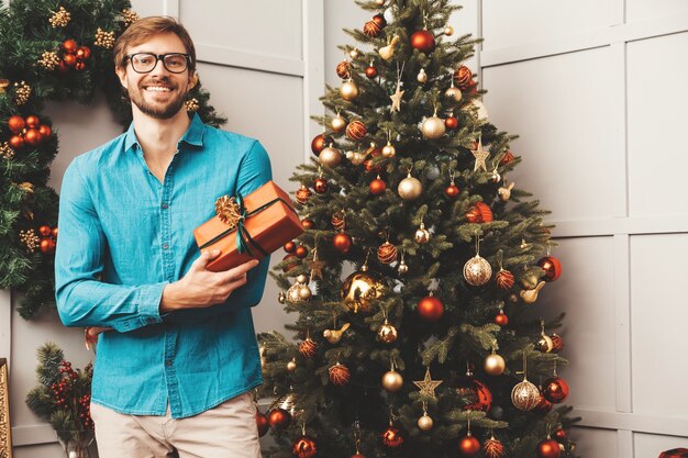 Портрет улыбающегося красавца, держащего подарок. Сексуальный бородатый мужчина позирует возле елки с настоящим.