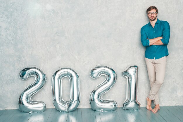 Портрет улыбающегося красавца, позирующего у стены. Сексуальный бородатый мужчина стоит возле Серебряных воздушных шаров 2021 года. С Новым 2021 годом. Металлические номера 2021