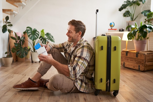 Foto gratuita ritratto di un bell'uomo sorridente seduto con una valigia sul pavimento con passaporto e biglietti aerei