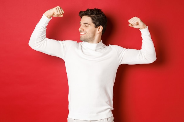 Portrait of smiling handsome man in white sweater flexing biceps and bragging with strength showoff ...