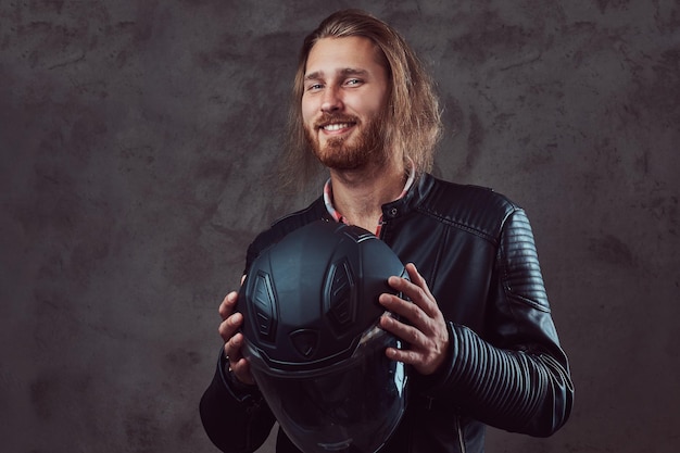 Portrait of a smiling handsome stylish redhead biker in a black leather jacket, holds motorcycle helmet, posing in a studio. Isolated on a dark background.