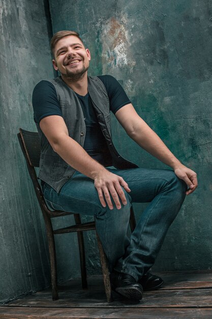 Портрет улыбающегося счастливого человека, сидящего на деревянном стуле на серой студии