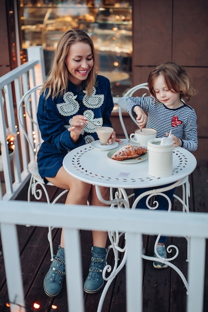 Ritratto di donna felice sorridente con capelli biondi in abito blu con anatre e stivali blu che si gode una tazza di caffè con sua figlia al bar. bella ragazza che mescola cacao seduta dalla madre a tavola all'aperto.