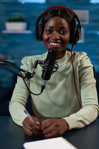 Portrait of smiling infuencer wearing headpones talking into professional microphone answering subscribers question during live stream. Social media vlogger recording video content. On air talk show