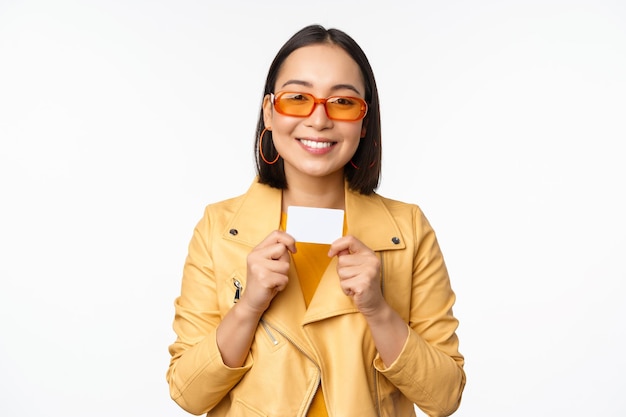 Portrait of smiling korean female model in sunglasses showing credit card standing over white background